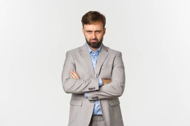 Kederli, kasvetli, gri takım elbiseli, kollarını göğsünde kavuşturmuş, beyaz arka planda poz veren, birine kızgın bir adamın portresi.