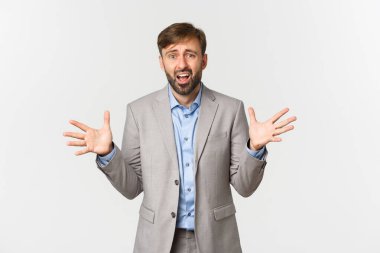Sakallı, gri takım elbiseli, ellerini yana açıp gergin görünen umutsuz ve endişeli iş adamının portresi.