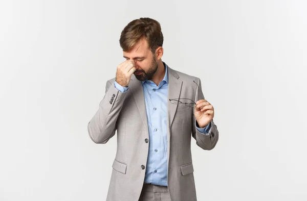 Retrato de empresário masculino cansado em terno cinza, óculos de decolagem e olhos esfregando, em pé sobre fundo branco — Fotografia de Stock