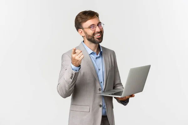 Portret van blije en tevreden zakenman in grijs pak en bril, werkend met laptop en wijzende vinger naar camera, prijzend goed idee, staande over witte achtergrond — Stockfoto