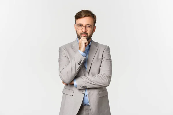 Portrait of thoughtful bearded businessman in glasses and grey suit, making his decision, squinting and looking at you, thinking while standing against white background — Stock Photo, Image