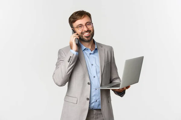 Beeld van een succesvolle en zelfverzekerde zakenman in een bril en een grijs pak, die telefoneert en een laptop vasthoudt, blij glimlacht op de camera, over een witte achtergrond staat — Stockfoto