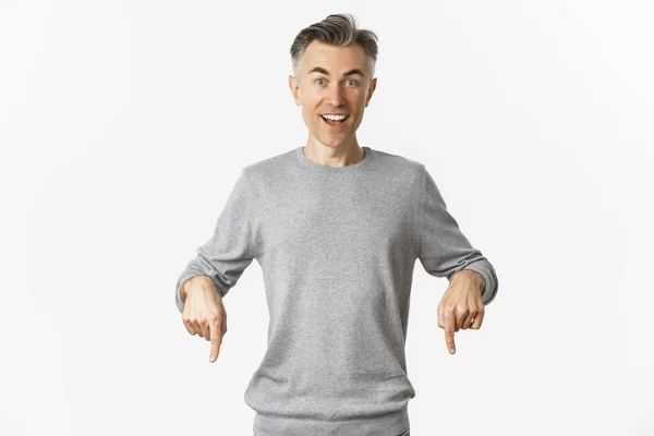 Imagem de homem de meia-idade bonito surpreso apontando os dedos para baixo, sorrindo e olhando curioso, de pé sobre fundo branco em camisola cinza — Fotografia de Stock