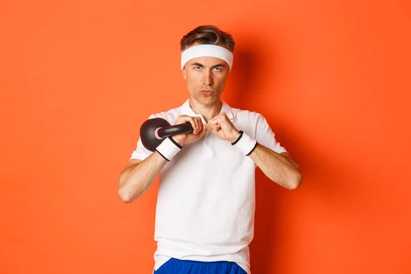 Konzept aus Workout, Fitness und Lifestyle. Nahaufnahme eines entschlossenen Fitness-Typen mittleren Alters, der Sport mit der Kettlebell macht, auf orangefarbenem Hintergrund stehend — Stockfoto