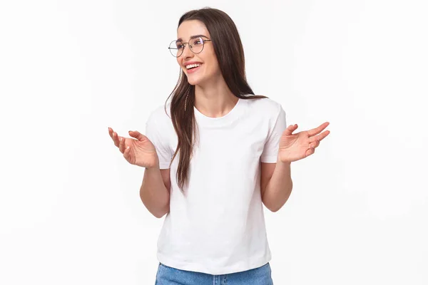 Waist-up portrait of carefree smiling attractive girl in glasses, white t-shirt jeans, gesturing hands sideways and look away with pleased smile, talking to coworkers, introduce her project — Stock Photo, Image