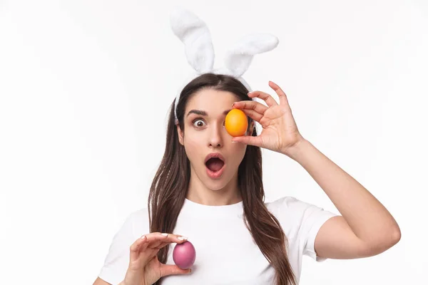 Pâques, vacances et concept de printemps. Portrait en gros plan d'une jeune femme amusée et enthousiaste aux oreilles de lapin, caméra à la bouche ouverte, tenant un œuf coloré au-dessus des yeux et à la main — Photo