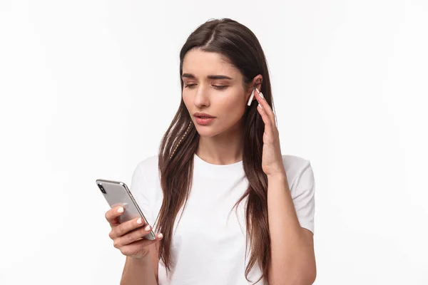 Close-up portrait of modern young woman in t-shirt, using wireless earphones listening music or podcasts, picking song in mobile phone, texting friend or calling person, white background — Stock Photo, Image