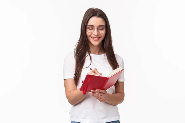 Portrait de sourire jolie fille caucasienne écrit dans son journal intime, préparer ses devoirs pour les cours d'anglais, sourire, avoir l'inspiration pour écrire des plans pour la semaine prochaine dans le planificateur, fond blanc — Photo