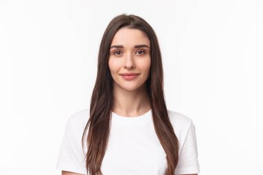 Güzel genç bir kadının yakın plan portresi, uzun kestane rengi saçlı ve sivilcesiz modern bir kız, ayakta duran basit bir gömlek ve gülümseyen bir kamera, olumlu arkadaşça bir ifade, beyaz bir arka plan.