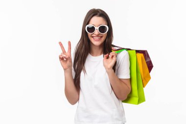 Alışveriş, eğlence ve yaşam tarzı konsepti. Rahat, kaygısız, çekici, modern, bayan, çantaları omuzlarının arkasında tutan, barış işareti yapan, güneş gözlüğü takan, yaz için kıyafet alan.