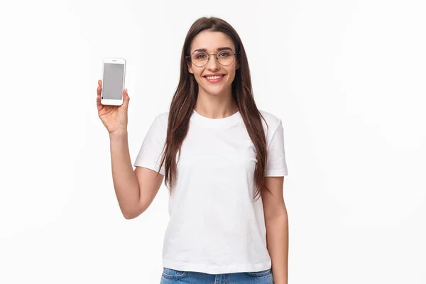 Технология и образ жизни. Портрет счастливой улыбающейся дружелюбной девушки в очках, футболке, с дисплеем смартфона, приложением для мобильного телефона, рекомендую загрузить его — стоковое фото