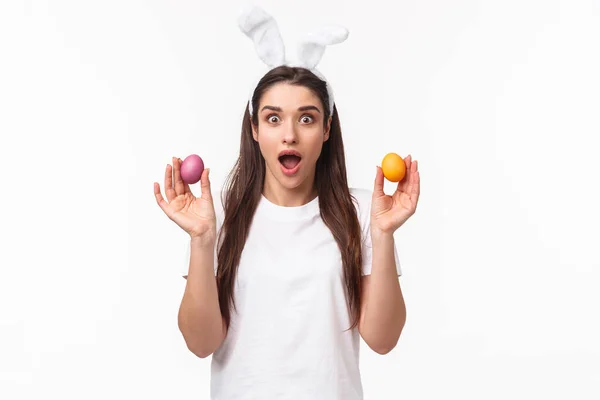 Portrait d'une jeune fille brune surprise et étonnée tenant deux œufs colorés, portant des oreilles de lapin, tenue de fête comme célébrant le jour de Pâques, profitant de vacances saintes, fond blanc — Photo