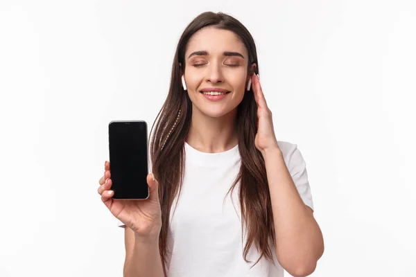 Close-up portrait of delighted, romantic young woman feeling satisfaction of listening favorite playlist, showing mobile phone screen, close eyes smiling and wearing wireless headphones — Stock Photo, Image