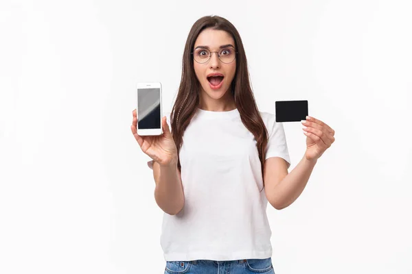 Porträt einer aufgeregten und glücklichen jungen Frau stellt neue Anwendung vor, bevorzugter Online-Shop, um Kleidung zu kaufen, Handy und Kreditkarte zu zeigen, amüsiert lächelnd, weißer Hintergrund — Stockfoto