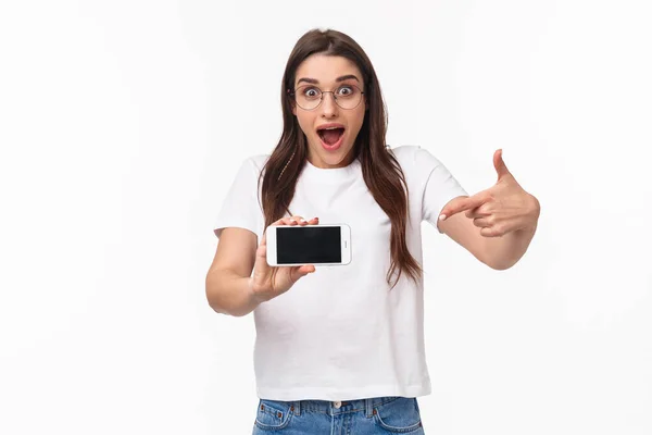 Comunicação, tecnologia e conceito de estilo de vida. Retrato de mulher jovem surpreso e animado em óculos apressar-se com o anúncio de incrível nova aplicação, apontando para o telefone móvel — Fotografia de Stock