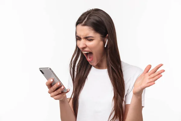 Close-up portrait of pissed-off aggressive and angry young brunette woman yelling at person while having phone call in wireless earphones, screaming at smartphone outraged — 图库照片