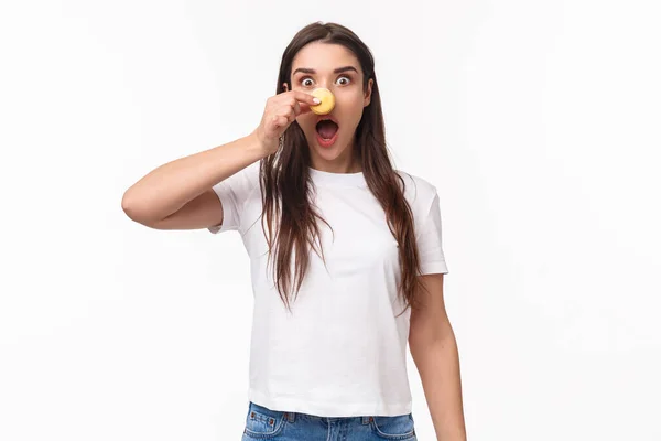Portrait de drôle adorable jeune femme brune en t-shirt, bouche ouverte excitée, air enthousiaste, blague faisant nez de macaron et appareil photo fixe, manger de délicieux desserts, fond blanc — Photo