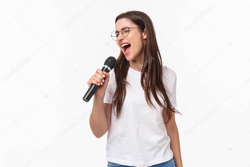 Carefree happy and relaxed young woman singing her heart out, performing song in karaoke, close eyes and place microphone near opened mouth, standing white background