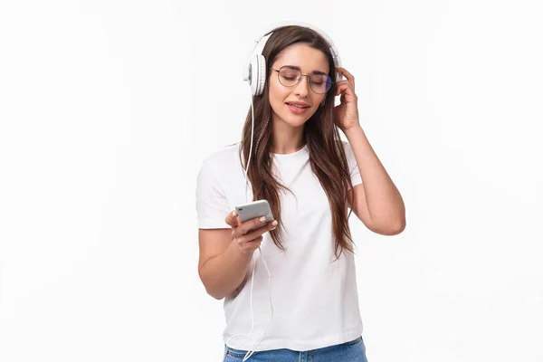 Technology, lifestyle and music concept. Portrait of pleased and relaxed young woman vibing and chilling as listening favorite song in headphones, hold mobile phone, smile and close eyes — Stock Photo, Image