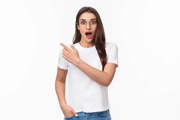 Waist-up portrait of enthusiastic, amazed and impressed young attractive girl in glasses talking about amazing new event or promo she saw online, pointing finger upper left corner, gasping astonished — Stock Photo, Image