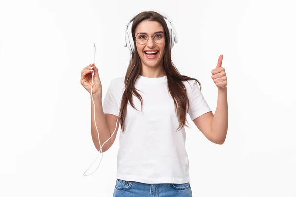 Waist-up portrait of excited, smiling happy and enthusiastic young girl in headphones, holding wire of earphones and show thumbs-up, ready to plug in to smartphone and listen some music — Stock Photo, Image