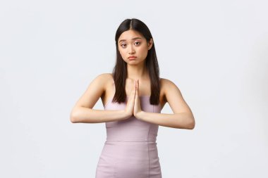 Lüks kadınlar, parti ve tatil konsepti. Akşam elbisesi içinde üzgün, Asyalı bir kız el ele tutuşmuş, yardım dileniyor, yardıma ihtiyacı var, yalvarıyor ya da özür diliyor, beyaz arka planda duruyor.