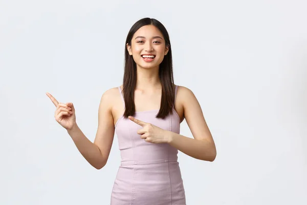 Concepto de celebración, belleza y glamour. Feliz sonrisa hermosa mujer asiática celebrando vacaciones en la fiesta en vestido de noche, señalando los dedos a la izquierda en la tienda con ropa hermosa, fondo blanco — Foto de Stock