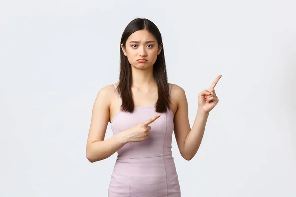 Luxury women, party and holidays concept. Upset and disappointed asian girlfriend pointing fingers upper right corner and frowning displeased, feeling upset about ruined celebration, white background — Stock Photo, Image