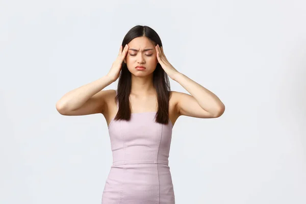 Mujeres de lujo, fiesta y concepto de vacaciones. angustiada joven asiática mujer en vestido de noche teniendo migraña, tocando la cabeza y mutilando de dolor, cerrar los ojos sufren dolor de cabeza, fondo blanco —  Fotos de Stock