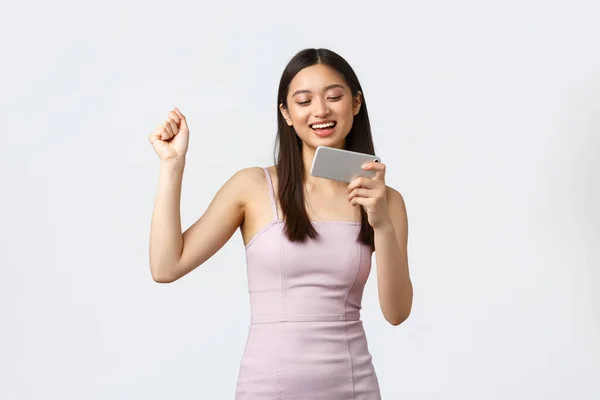 Mujeres de lujo, fiesta y concepto de vacaciones. Feliz mujer bonita satisfecha en vestido de noche, hacer danza campeón como ganar en el juego móvil, celebración de teléfono inteligente y bomba de puño emocionado, fondo blanco — Foto de Stock