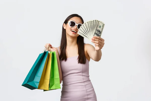 Style de vie, shopping et concept de tourisme. Sourire riche jeune femme coréenne en robe et lunettes de soleil, rire insouciant, acheter des vêtements, tenant de l'argent et des sacs à provisions, fond blanc — Photo