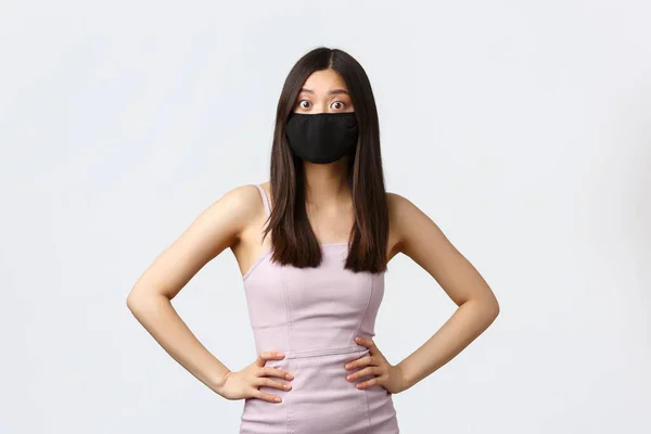 Covid-19, social distancing quarantine and leisure concept. Surprised and overwhelmed young asian woman in face mask, stare at camera amazed, wear face mask and evening dress, white background — Stock Photo, Image