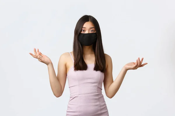 Covid-19, socialt distanserande karantän och fritidskoncept. Förvirrad asiatisk kvinna i aftonklänning och ansiktsmask, kan inte förstå vad som hände, rycka med händerna i sidled, se förbryllad ut — Stockfoto