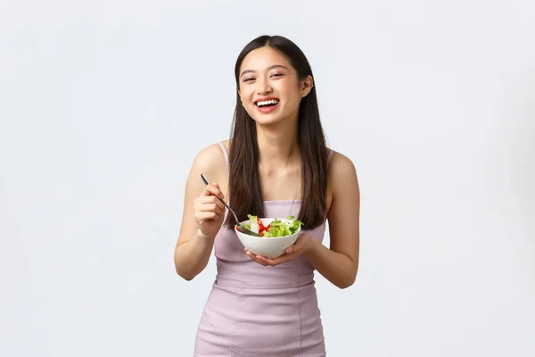 Estilo de vida saludable, ocio y personas concepto de emociones. alegre sonriente asiático chica comer ensalada y riendo feliz, vistiendo partido vestido, de pie blanco fondo — Foto de Stock