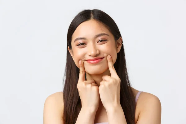 Conceito de beleza, moda e emoções das pessoas. Close-up retrato de adorável bobo asiático menina no vestido, puxando feliz sorriso com os dedos sobre os lábios, sentindo otimista e alegre, fundo branco — Fotografia de Stock