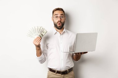 İş ve e-ticaret. Adam para geliri ile şaşkına dönmüş, internette çalışıyor, dizüstü bilgisayar kullanıyor, beyaz arka planda duruyor.