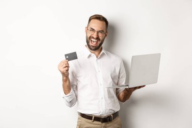 İnternetten alışveriş. Kredi kartı gösteren yakışıklı ve internetten sipariş vermek için dizüstü bilgisayar kullanan beyaz arka planda duran bir adam.