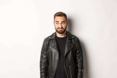 Siyah deri ceketli kararsız kasvetli adam, sola bakıyor ve kendini rahatsız hissediyor, beyaz arka planda duruyor.