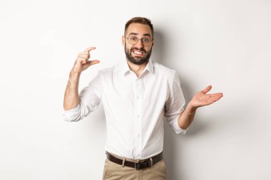 İşadamı küçük bir jest yapıyor, canı sıkkın görünüyor, omuz silkiyor, beyaz arka plana karşı duruyor.