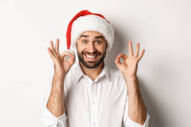 Parti, kış tatili ve kutlama konsepti. Neşeli adam Noel 'den zevk alıyor ve iyi bir işaret veriyor, gülümsüyor, Noel Baba şapkası takıyor, beyaz arka plan