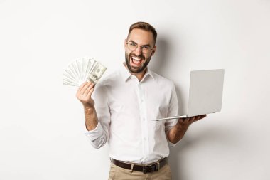 İş ve e-ticaret. Heyecanlı iş adamı elinde dolar ve laptop tutuyor, internette çalışıyor, beyaz arka planda duruyor.