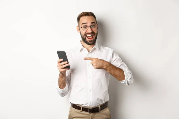 Homme regardant excité et pointant du doigt le téléphone mobile, montrant une bonne offre en ligne, debout sur fond blanc — Photo