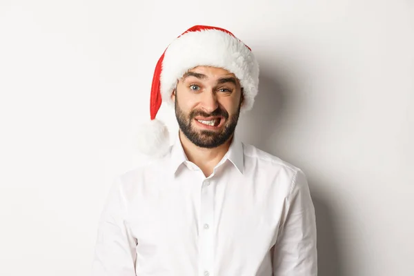 Gros plan d'un gars maladroit en chapeau de Père Noël s'excusant, se sentant mal à l'aise, debout sur fond blanc. Concept de Noël — Photo