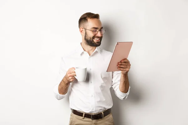 Knappe zakenman drinken van koffie en lezen op digitale tablet, glimlachend blij, staande over witte achtergrond — Stockfoto