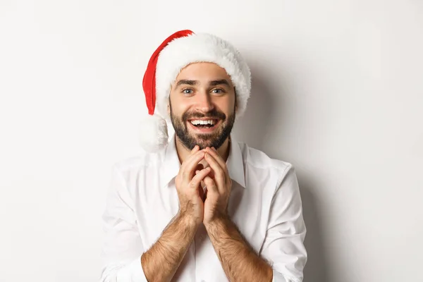 Party, Winterurlaub und Festkonzept. Aufgeregter und hoffnungsvoller Mann mit Weihnachtsmütze, der das Weihnachtsgeschenk mit Staunen betrachtet, weißer Hintergrund — Stockfoto