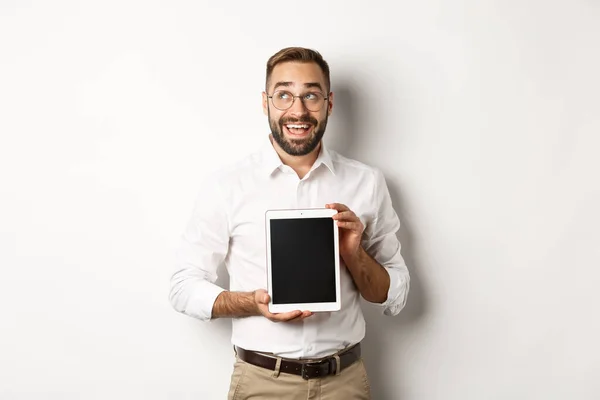 Shopping et technologie. Homme réfléchi montrant l'écran de la tablette numérique, regardant le coin supérieur gauche et pensant, debout sur fond blanc — Photo