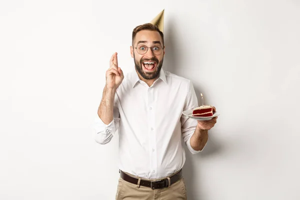 Holidays and celebration. Excited man having birthday party, making wish on b-day cake and cross fingers for good luck, standing against white background — Stock Photo, Image