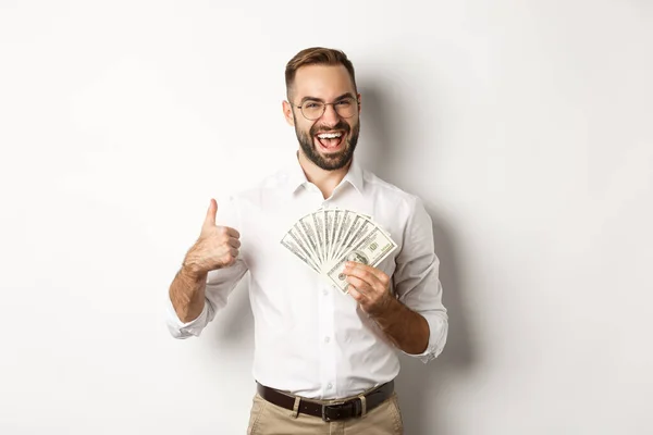 Entusiasmado homem rico segurando dinheiro, mostrando polegar para cima na aprovação, de pé sobre fundo branco — Fotografia de Stock
