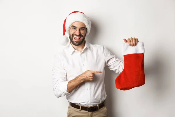 Szczęśliwy człowiek w czapce Świętego Mikołaja świętuje zimowe wakacje, wskazując na skarpetkę świąteczną i uśmiechnięty, trzymając prezenty, stojąc nad białym tle — Zdjęcie stockowe