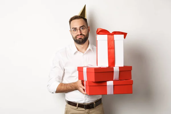 Праздники и праздники. Недовольный парень держит подарки на день рождения и выглядит разочарованным, не любит подарки, стоя на белом фоне — стоковое фото